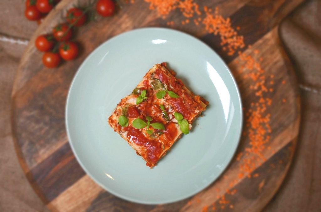 Tanier s lasagnami na drevenom stole s rozsypanými ingredienciami ako paradajky a červená šošovica.