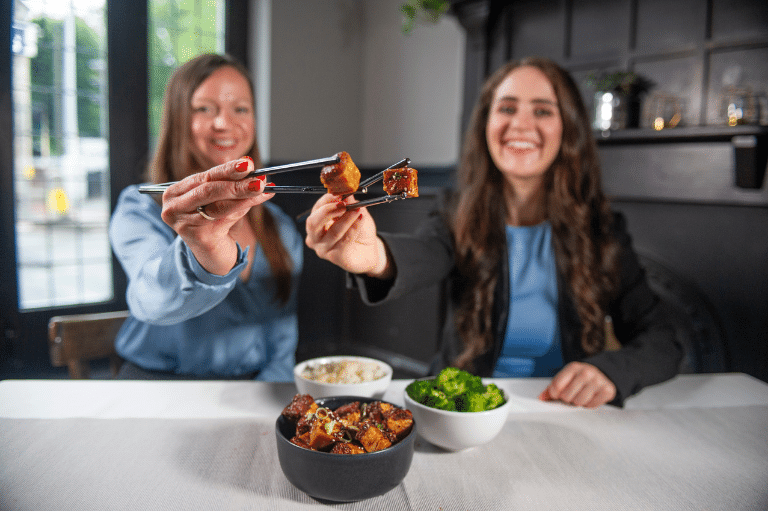 Dve ženy, sediace za prestretým stolom, držia v paličkách na jedenie kultivované bravčové mäso od firmy Higher Steaks.